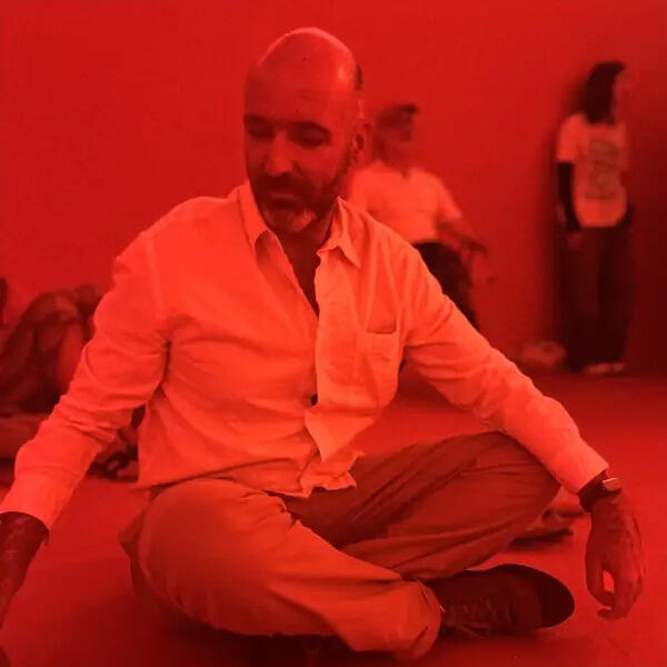 João Soares sentado no chão em postura de meditação, numa sala iluminada a vermelho